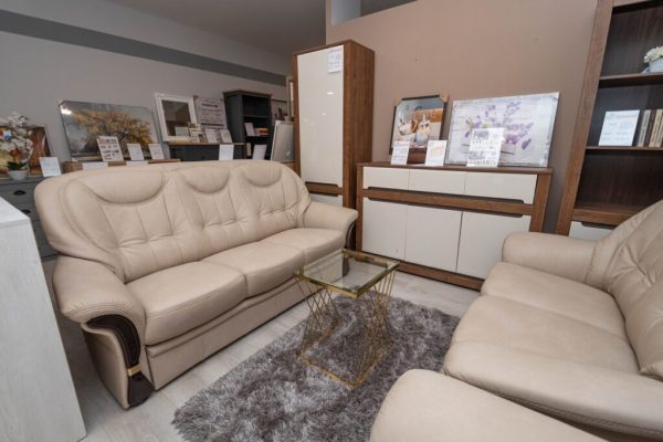 Egy modern nappaliban két egymással szemben álló, bézs bőrből készült BILBAO klasszikus & elegáns ülőgarnitúra kanapé látható, szürke shag szőnyegen üveg dohányzóasztallal, amelyet a háttérben szekrények és bekeretezett művészeti alkotások egészítenek ki.