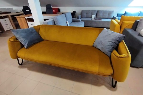 A FERDINAND design kanapé, egy mustársárga kanapé fekete fémlábakkal és két szürke párnával, szürke kanapék és fabútorok mellett látható egy bemutatóteremben.
